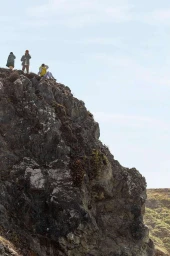 Three figures standing and sitting atop a rocky cliff edge under an open overcast sky, photographed by Andre L Perry.