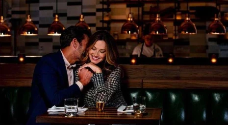 A man in a dark blue blazer whispering to a smiling woman in a patterned top at a restaurant table.