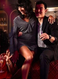 Smiling woman in a grey metallic dress sitting on a man's lap holding a champagne glass inside a dimly lit bar.