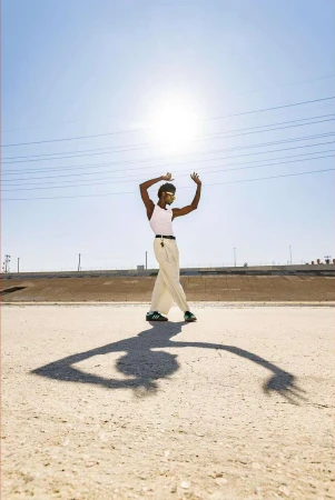 Black man in white tank top and cream trousers dancing outdoors beneath bright overhead sun with long dramatic shadow on ground