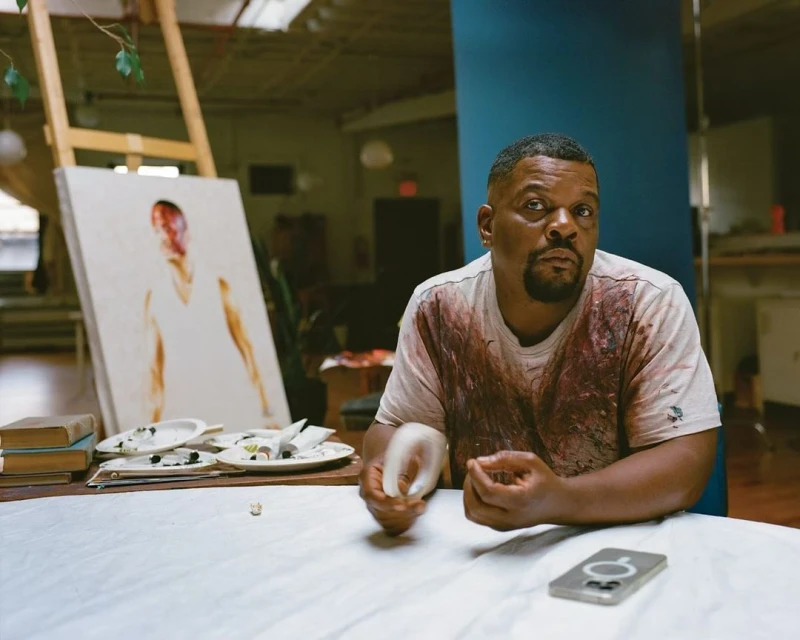 Artist sits in studio surrounded by supplies, canvas art on easel, wearing paint-splattered shirt, holding sculpting tools, serious expression.