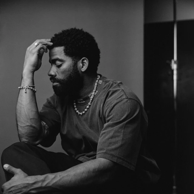 Monochrome portrait of a pensive adult male with a beard, wearing jewelry and casual attire, seated indoors in a studio setting.