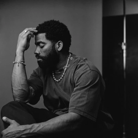Monochrome portrait of a pensive adult male with a beard, wearing jewelry and casual attire, seated indoors in a studio setting.
