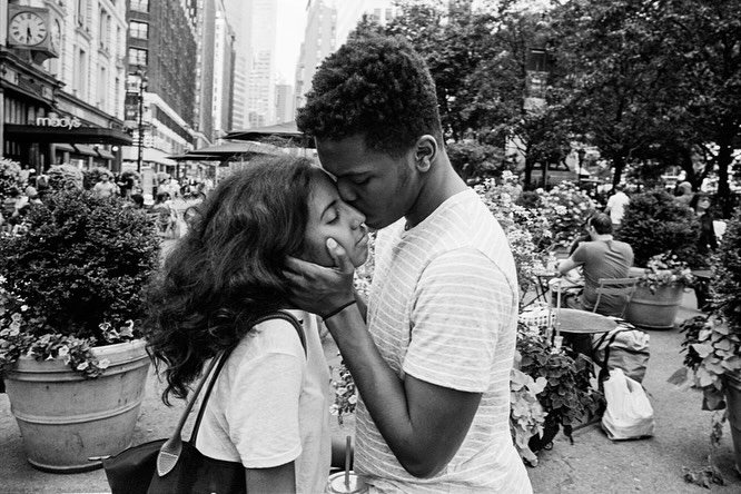 A young couple in an affectionate embrace, the man kissing the woman's forehead, set against an urban backdrop with city buildings and trees.