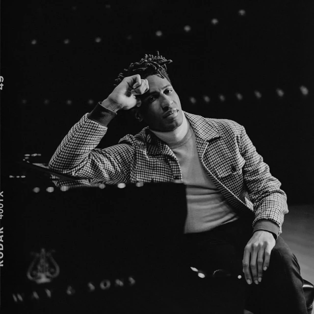 Black and white portrait of a stylish adult male in a patterned jacket, posing with his hand on a grand piano in a dimly lit studio.