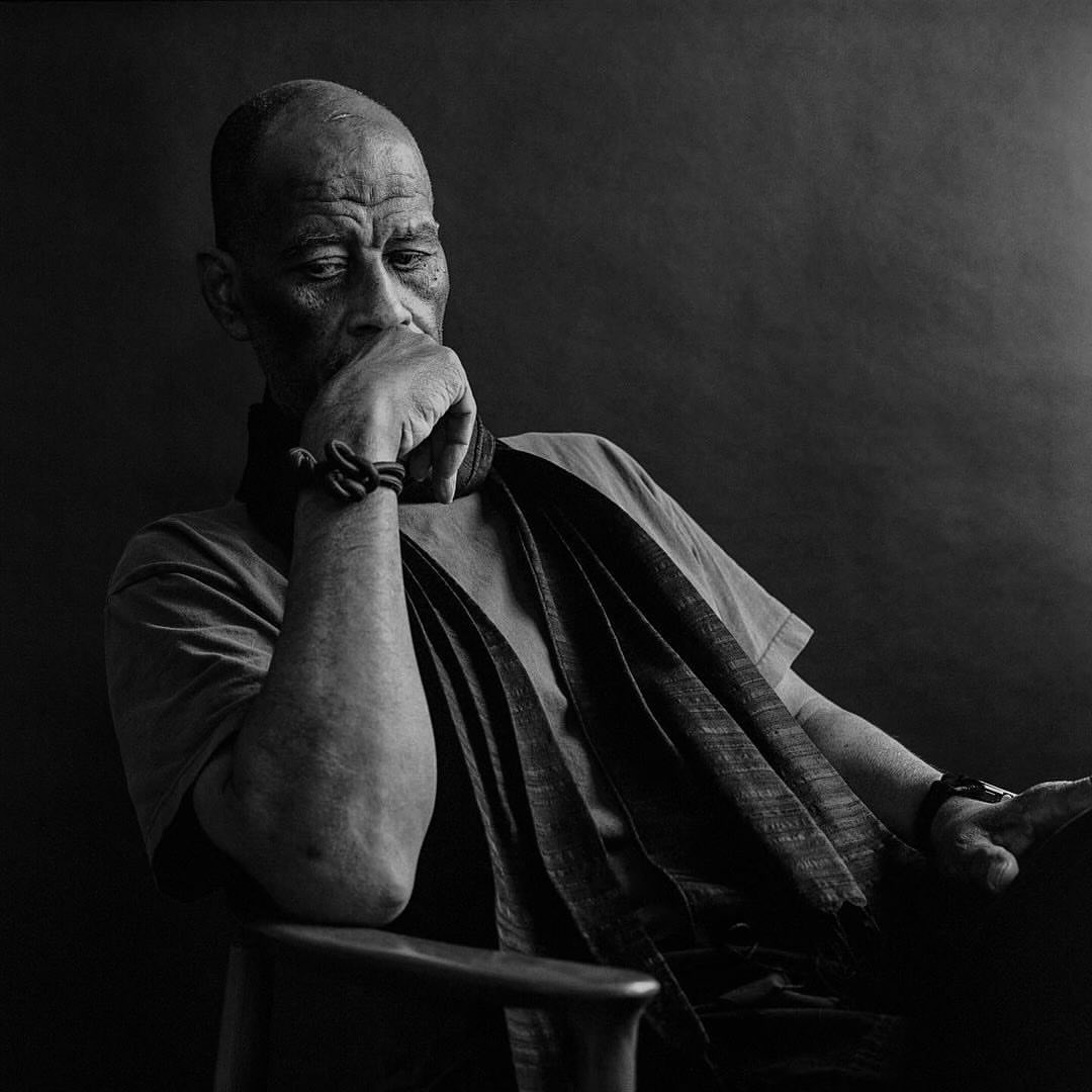 Black and white portrait of an elderly man with a pensive expression, wearing a scarf, sitting in a chair against a dark backdrop.