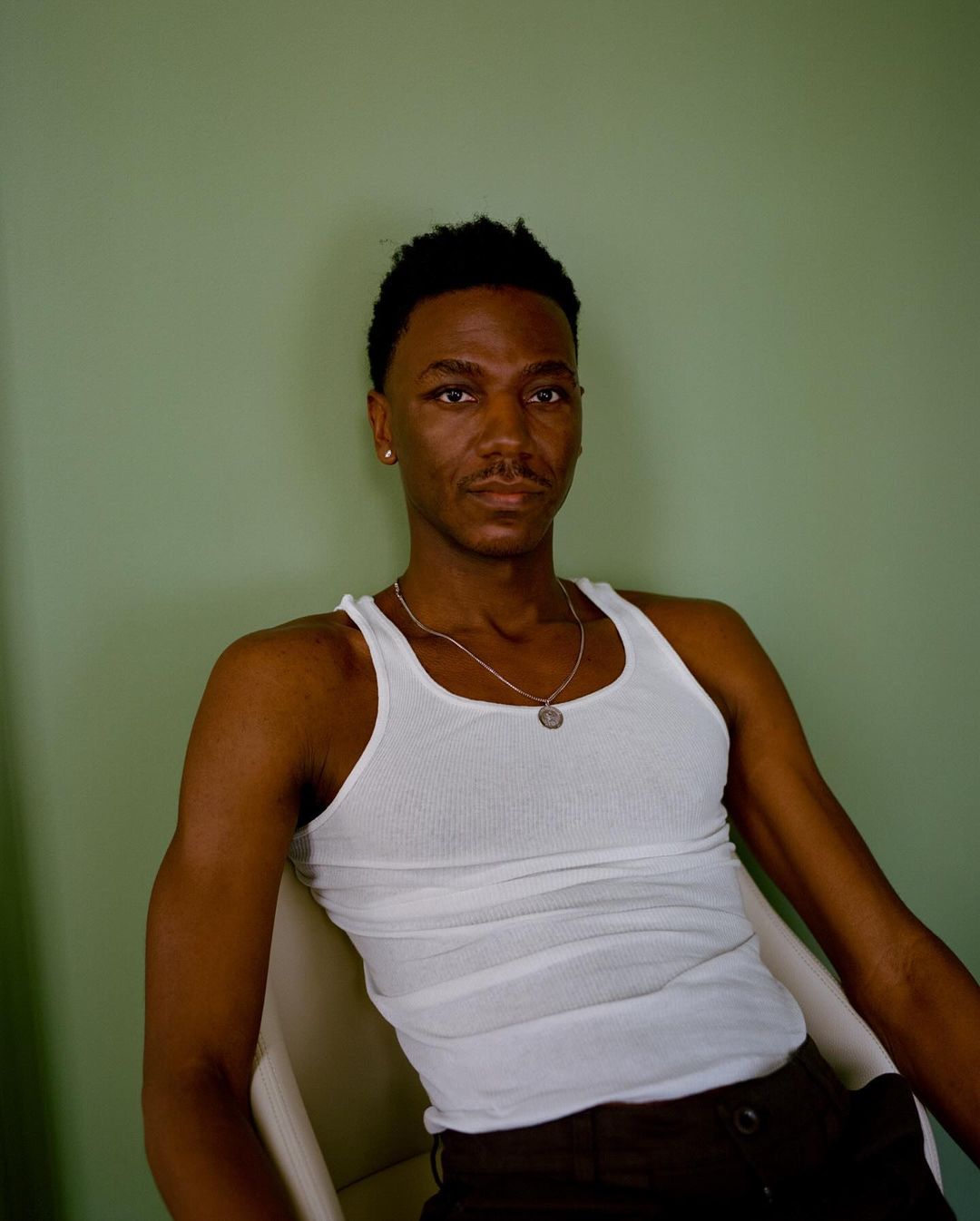 Portrait of a Black man wearing a white tank top and a silver necklace seated against a plain green wall.