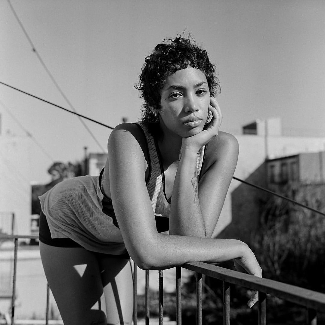 Black and white portrait of a woman in a tank top leaning on a railing outside with buildings and sky in the background.