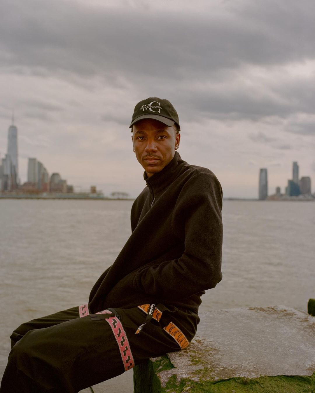 Young man poses near water with city skyline in the background. He wears a baseball cap, black top, and pants with striped details.