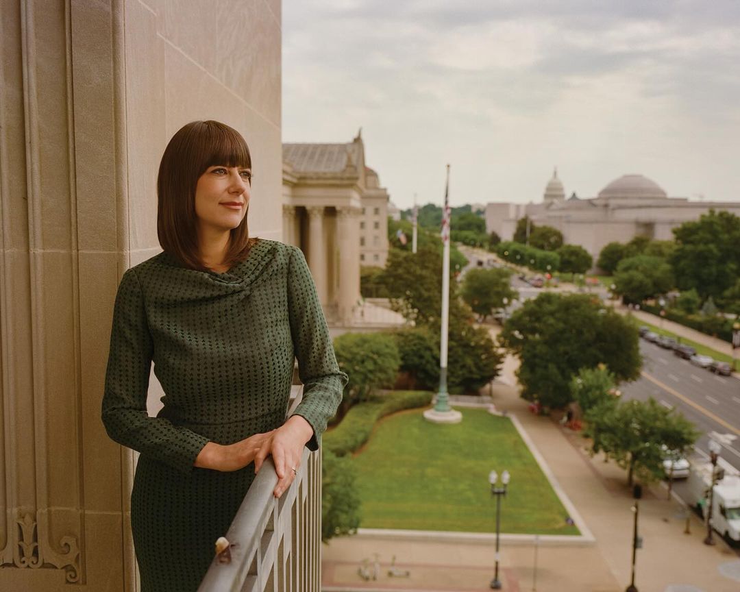 Woman in green dress poses on balcony overlooking Washington DC buildings, greenery, and street. Cloudy sky above in daytime.