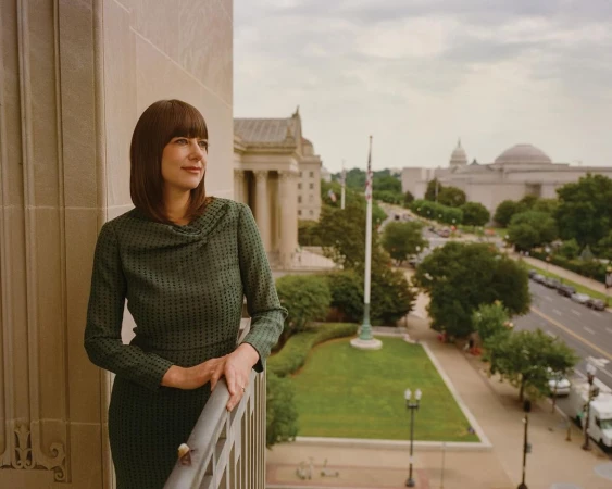 Woman in green dress poses on balcony overlooking Washington DC buildings, greenery, and street. Cloudy sky above in daytime.