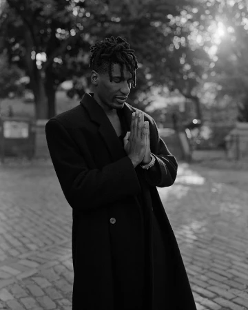 Black and white portrait of a black man wearing a dark coat with hands clasped in prayer outdoors in New York by Andre Wagner