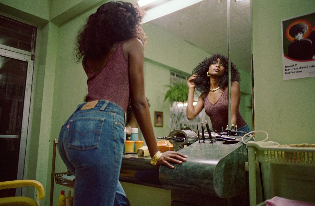 A young woman with dark curly hair and blue jeans poses in a vintage-style indoor beauty salon in front of a mirror.