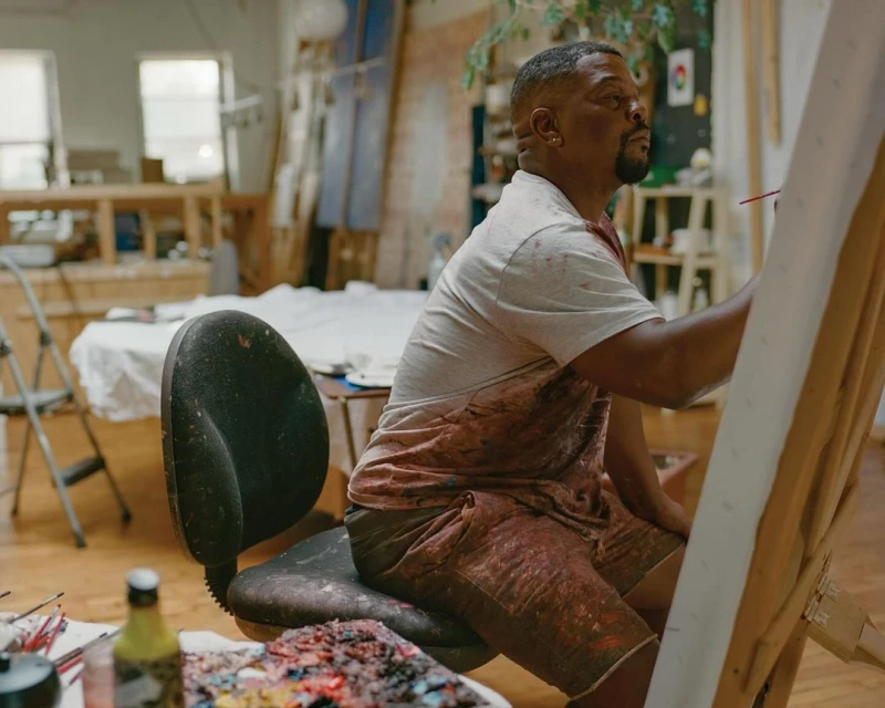 Artist at work in art studio, painting on canvas. African American male wearing paint-splattered clothes, art supplies visible in foreground.