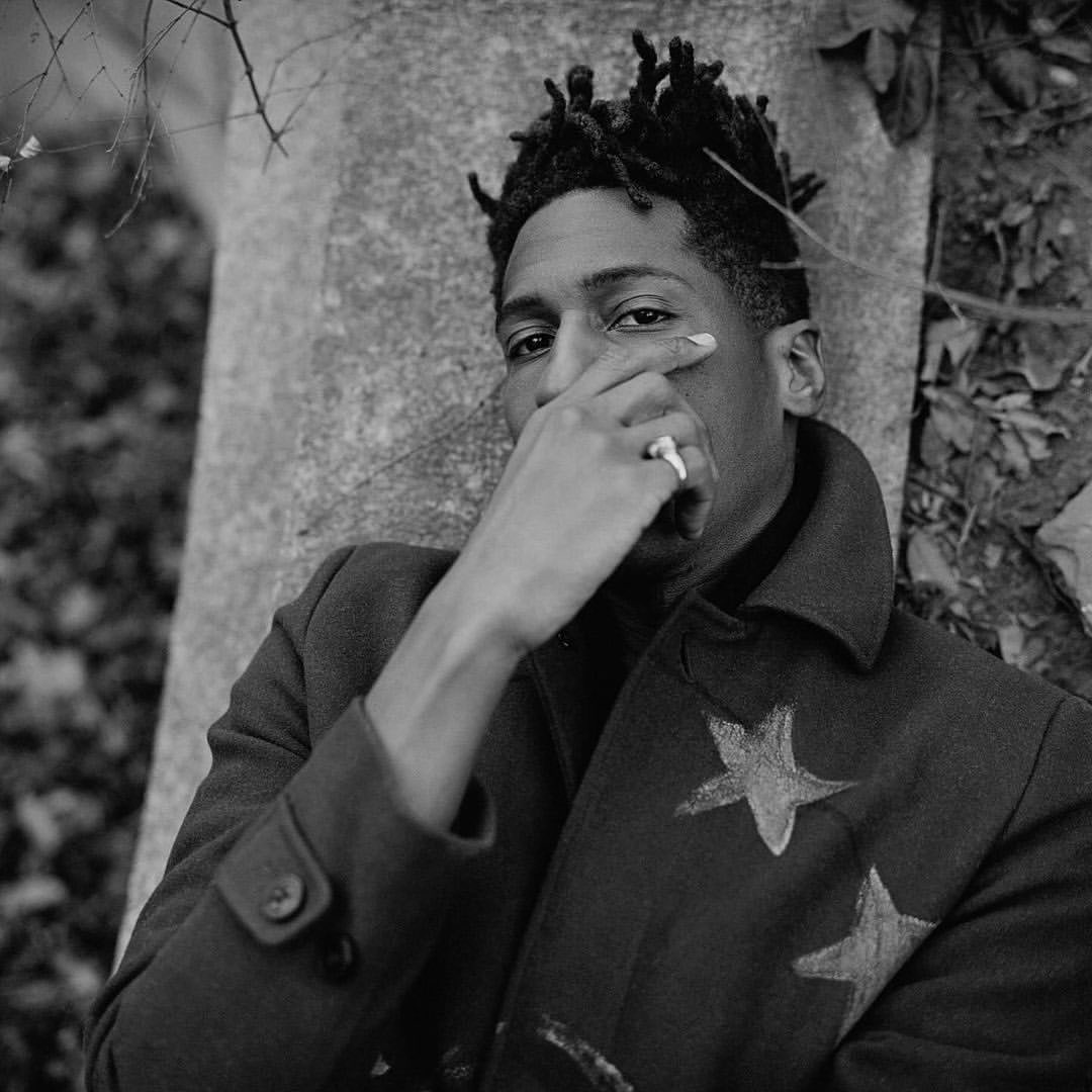 Black and white portrait of a Black male leaning against a tree, wearing a coat with stars, against a natural background.