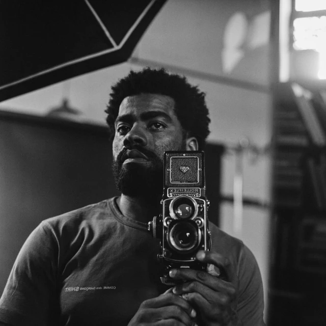Black and white portrait of an adult man holding a vintage twin-lens reflex camera in a photo studio with softbox lighting.
