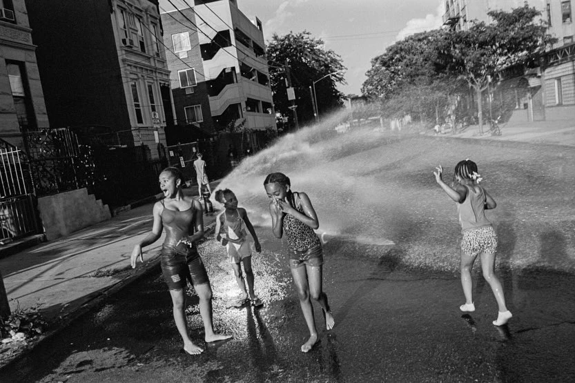A group of children play under a spray of water from a fire hydrant on a city street, on a hot day. Black and white street photography.