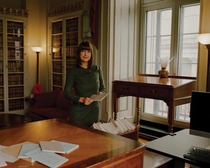Woman in green dress stands in traditional office with books, papers, and wooden furniture, holding papers in front of a window.