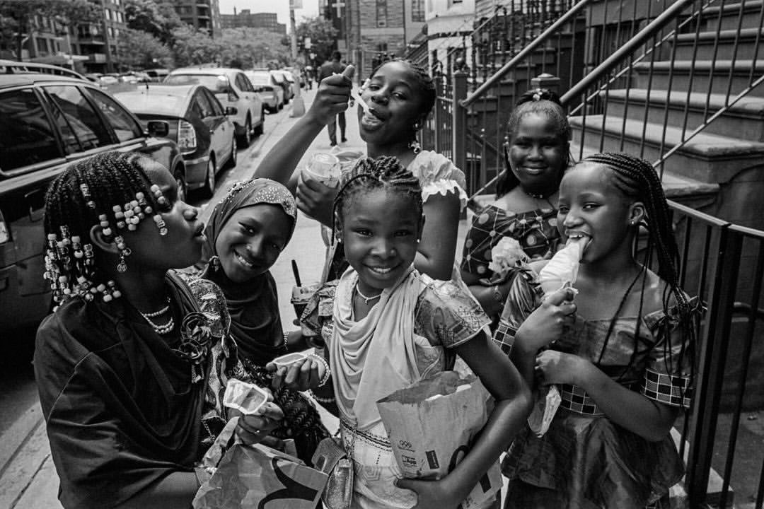 A monochrome street scene captures six young African girls enjoying frozen treats on a sidewalk in front of a brownstone building.