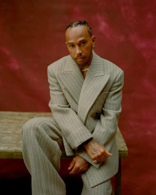 Portrait of Lewis Hamilton wearing a tan pinstripe suit, seated on a wooden surface against a mottled maroon backdrop, studio lighting.