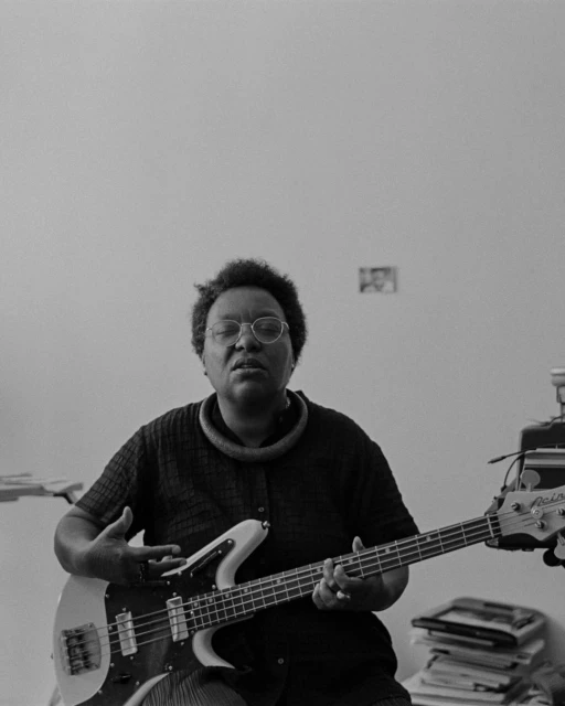 Black and white portrait of an adult woman playing an electric bass guitar, wearing a necklace, and eyeglasses indoors.