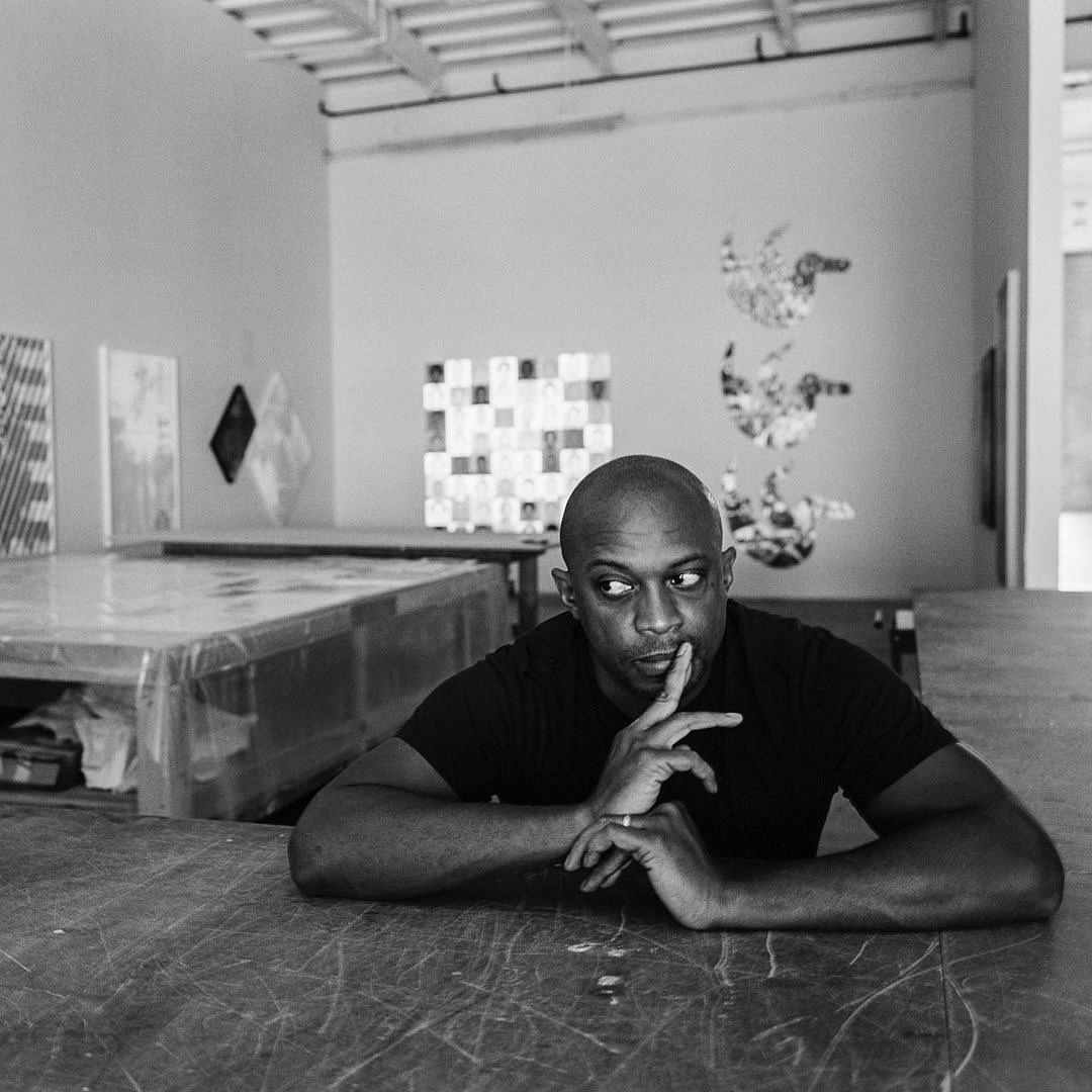 Monochrome portrait of a bald Black man in a black t-shirt resting his chin on his fingers while seated at a worn wooden table.