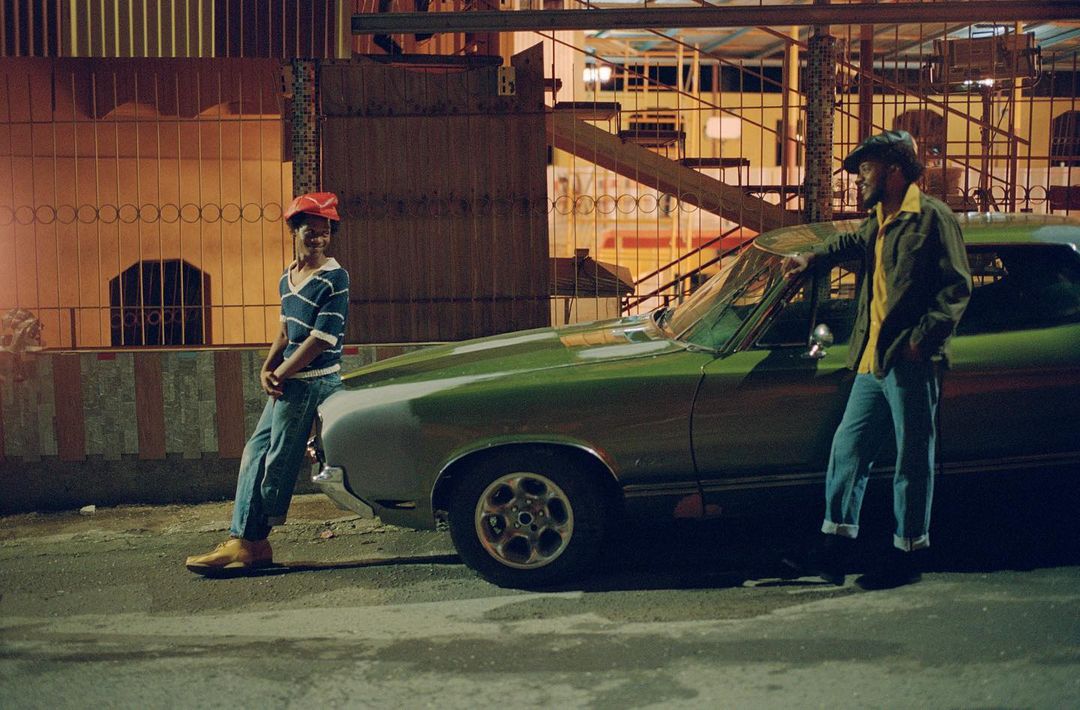 Two men posing by a vintage green car outdoors at night. One man leans against the car, another stands with a red hat and striped shirt.