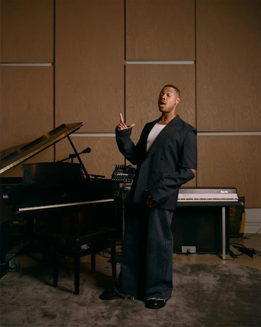 Adult man in an open black blazer and wide-leg pants stands next to a black piano and keyboard in an indoor music studio setting.