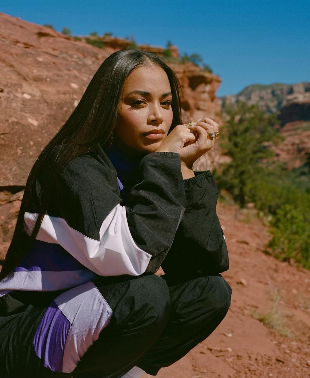 A young woman with dark hair kneels outside, wearing a black, white, and purple tracksuit, against a rocky, mountainous background.