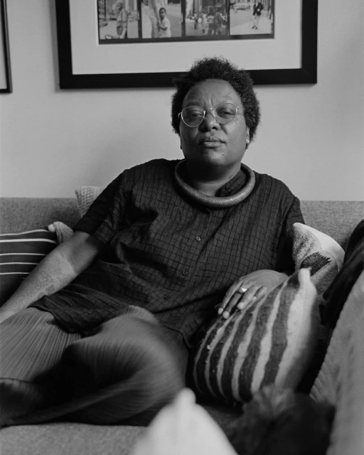 Black and white portrait of a woman with glasses, wearing a necklace, sitting on a couch with pillows and a framed picture on the wall.
