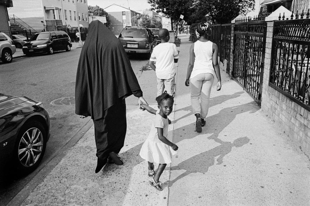 A black and white photograph shows an adult and child holding hands walking on a sidewalk, with cars and buildings in the background.