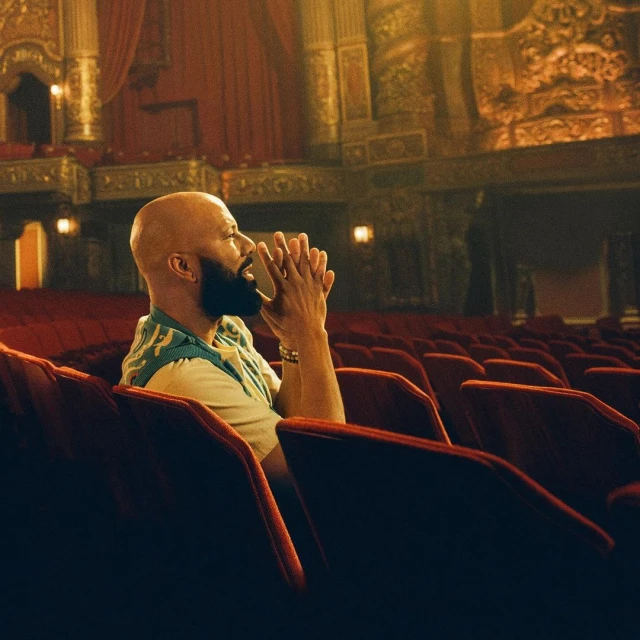 Man sitting in theatre seating, looking up with hands clasped. Warm lighting and deep red theatre seating fill the scene.