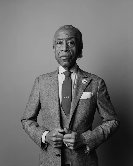 Black and white portrait of an adult male in a grey suit and red tie posing in front of a grey background in a photo studio.