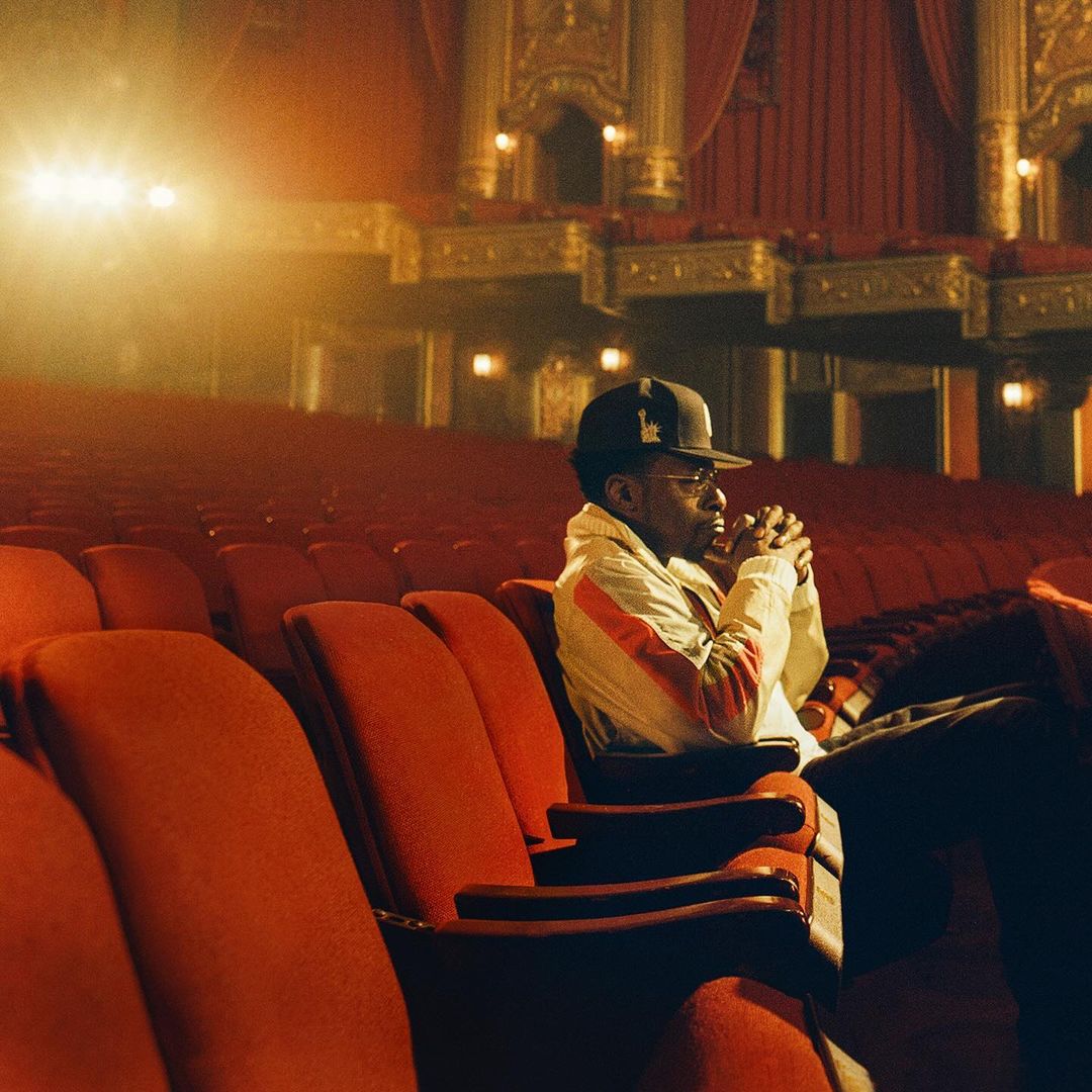 Man in baseball cap and jacket sits alone in a red theater seat. The theater's architecture is visible in the background.