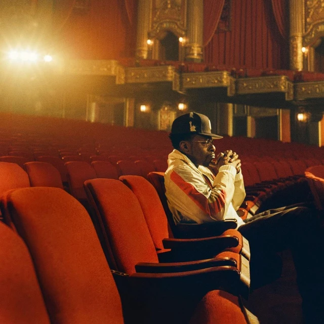 Man in baseball cap and jacket sits alone in a red theater seat. The theater's architecture is visible in the background.