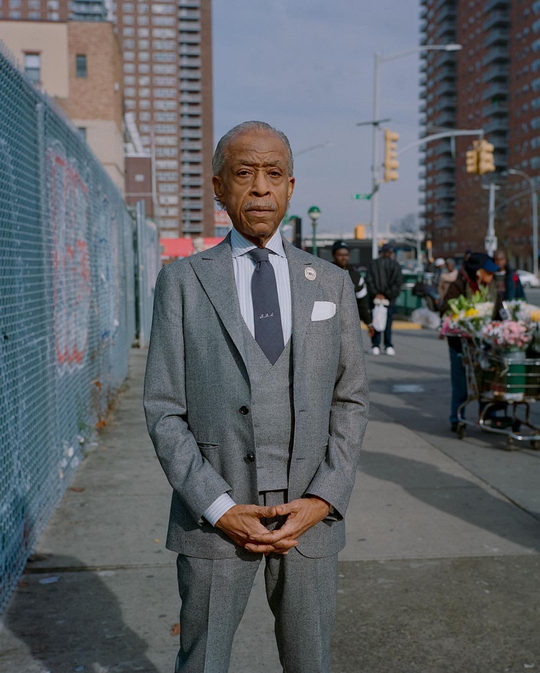 Portrait of a well-dressed older man in a grey suit, standing outdoors on a city street with buildings and people in the background.
