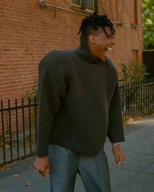 Portrait of an adult man with dreadlocks wearing a gray turtleneck sweater and gray pants standing in front of a brick wall.