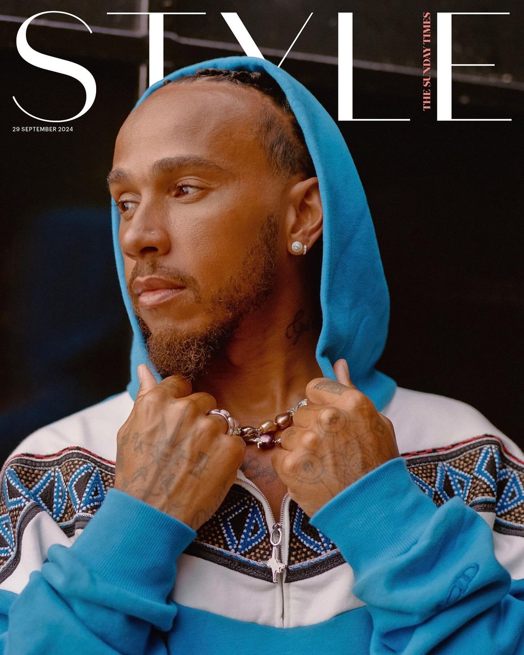 Lewis Hamilton, adorned with jewelry and a hoodie, gazes off-camera on a magazine cover, set against a blurred dark backdrop.