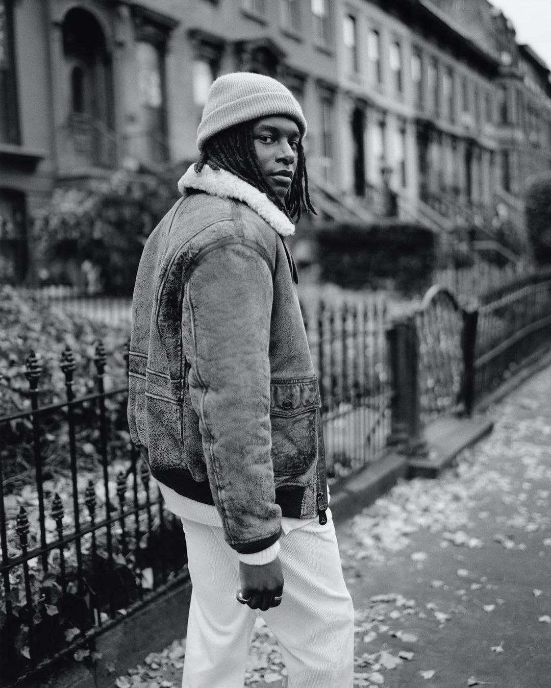 A person in a beanie and shearling jacket looks over their shoulder on a city street with brick buildings and fallen leaves.