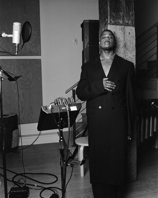 Black and white portrait of an African-American man in a dark coat standing in a music studio with a microphone and audio equipment.