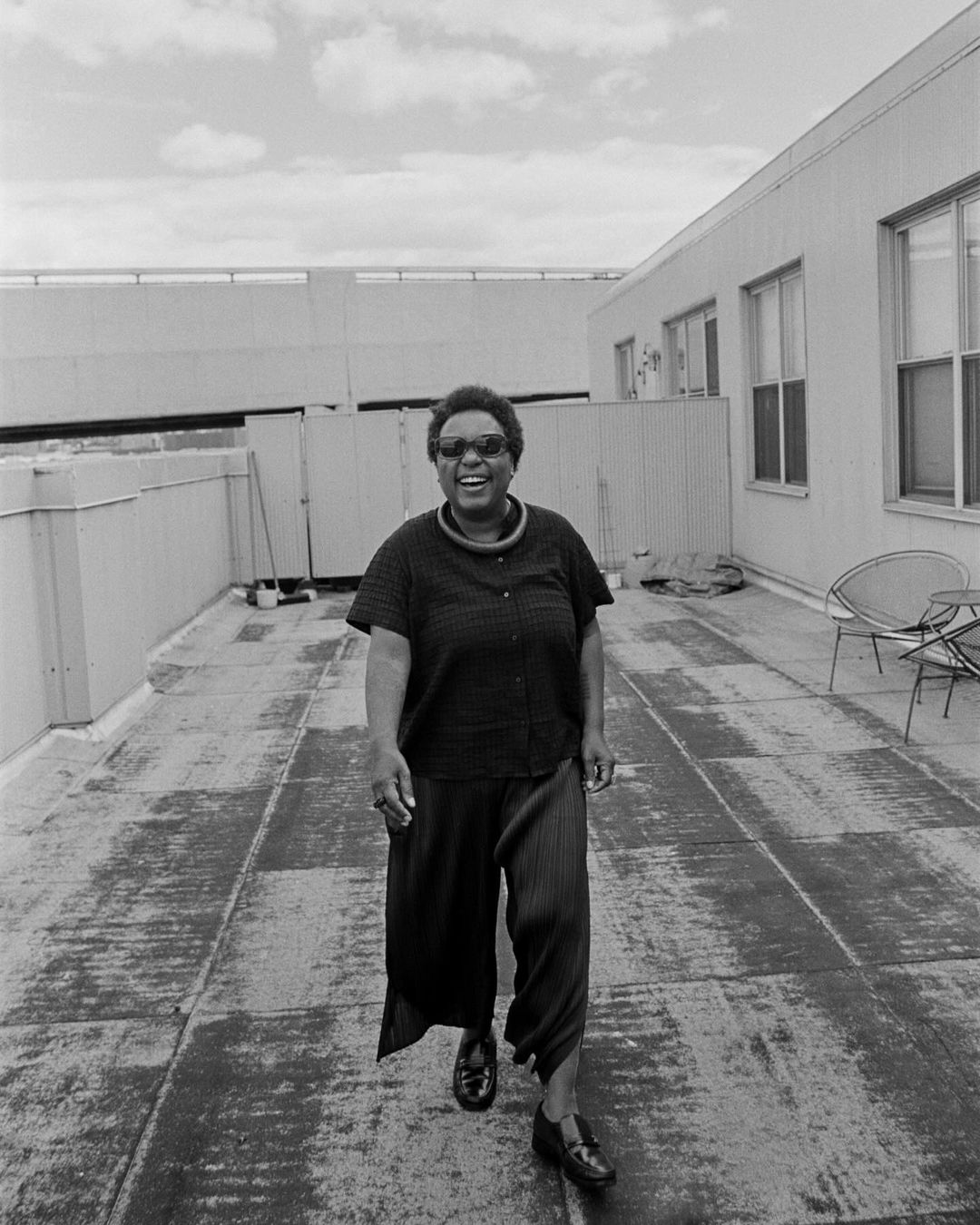 Black and white medium shot of an adult woman smiling and walking towards the camera on a rooftop on a sunny day.