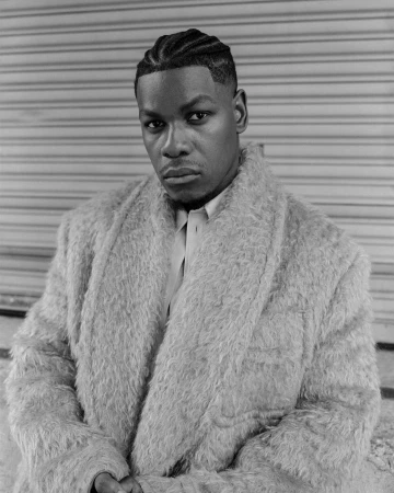 Portrait of John Boyega wearing a textured fluffy coat and braided hairstyle against a rolling shutter door by Andre Wagner