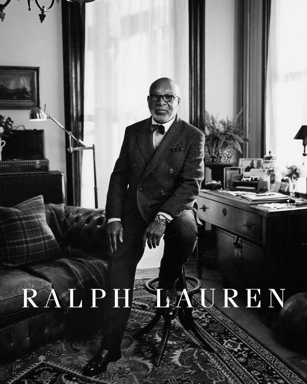 Black and white portrait of an African American man wearing a suit and bow tie, seated on a stool in a room with traditional decor.