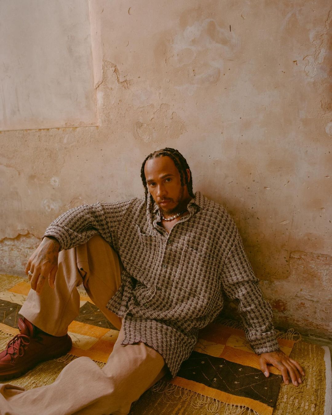 Man sits on patterned rug in neutral tones. He wears a patterned shirt and tan pants, and is set against a textured wall.