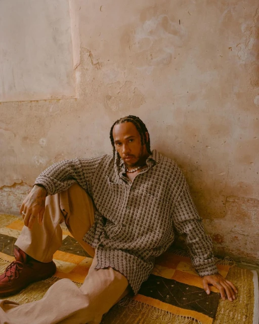 Man sits on patterned rug in neutral tones. He wears a patterned shirt and tan pants, and is set against a textured wall.