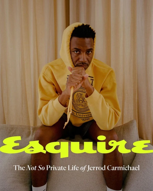 Man in a yellow hoodie sitting on a grey couch with the magazine 'Esquire' visible in the foreground in an indoor setting.