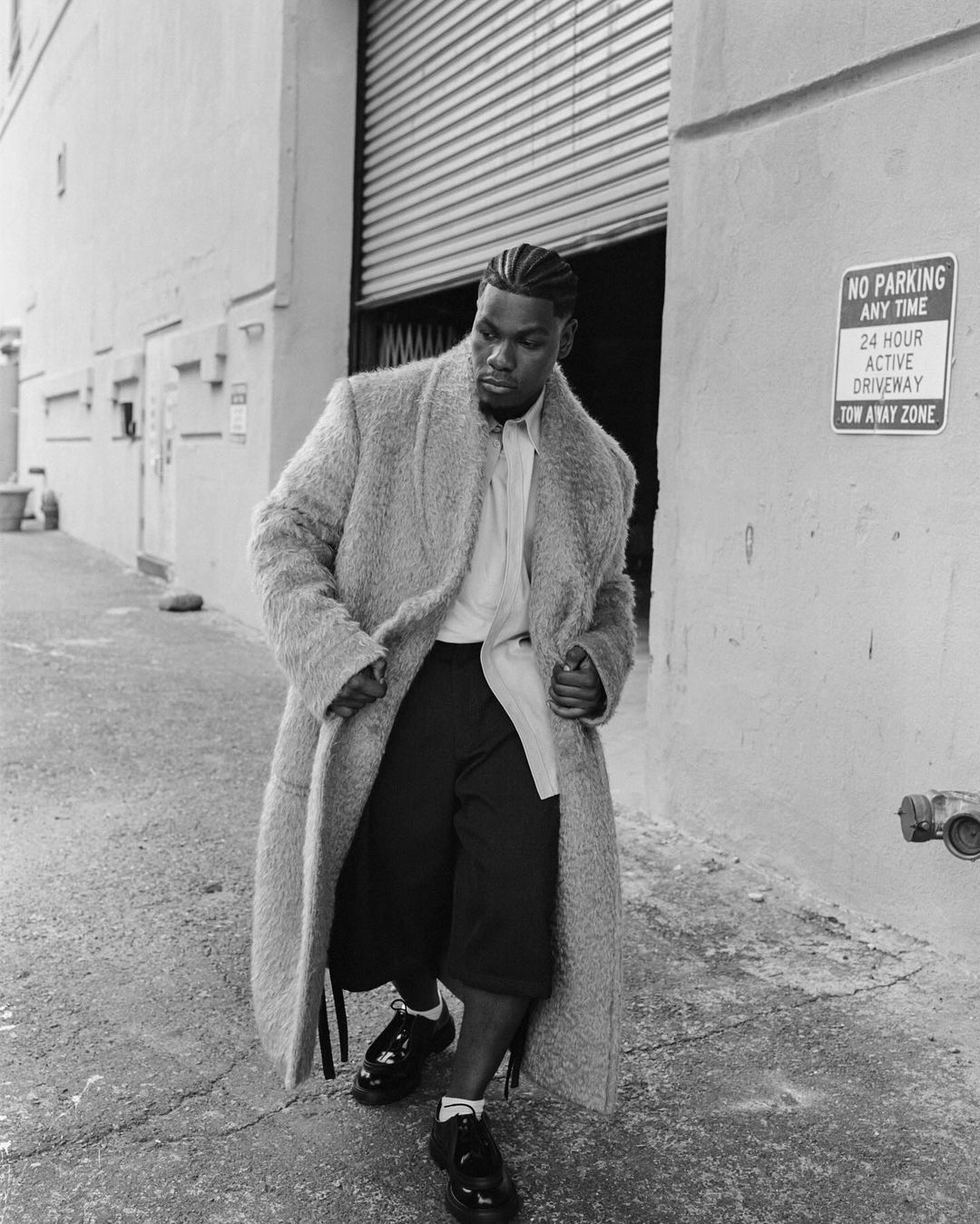 Monochrome shot of an adult male in an overcoat, culottes, and dress shoes standing in an alley next to a metal door.