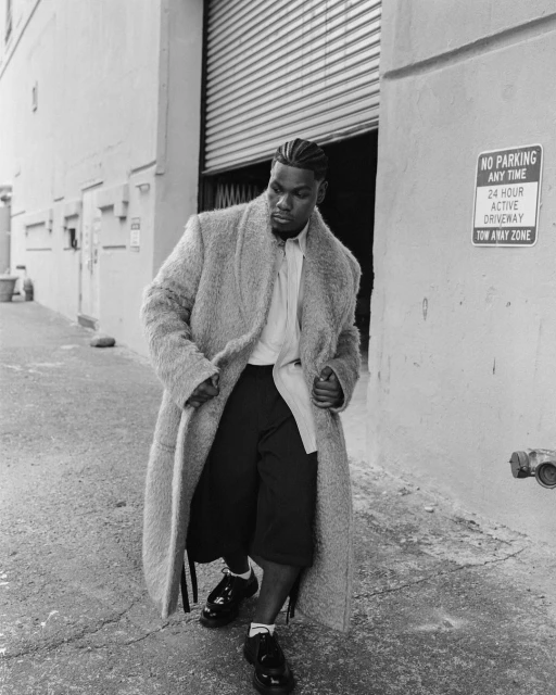 Monochrome shot of an adult male in an overcoat, culottes, and dress shoes standing in an alley next to a metal door.