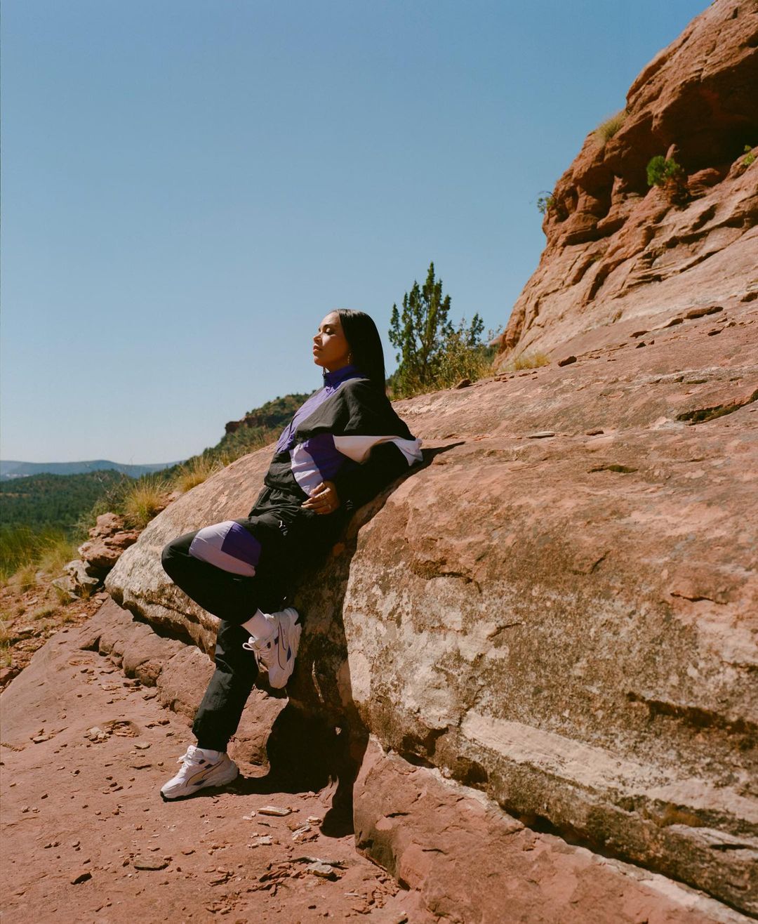 A young woman in a patterned tracksuit sits on red rocks against a clear blue sky, blending fashion with the natural desert landscape.