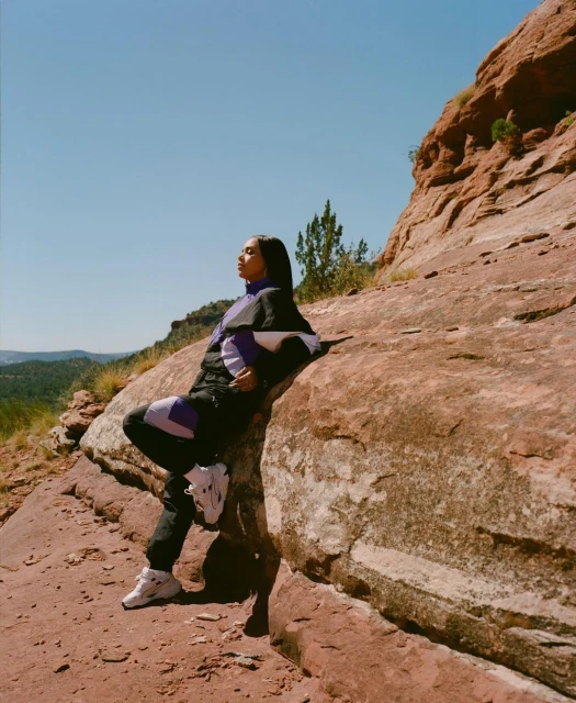 A young woman in a patterned tracksuit sits on red rocks against a clear blue sky, blending fashion with the natural desert landscape.